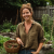 Willow Greene smiling in her backyard garden, holding a basket of fresh herbs and vegetables beside raised garden beds.